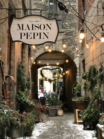 A wide brick alleyway with glowing holiday lights, green pine garlands, and a generally festive atmosphere.