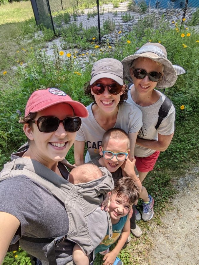 A top-down long-arm selfie of two young women, one middle-aged woman, two young kids, and a baby in a front pack.